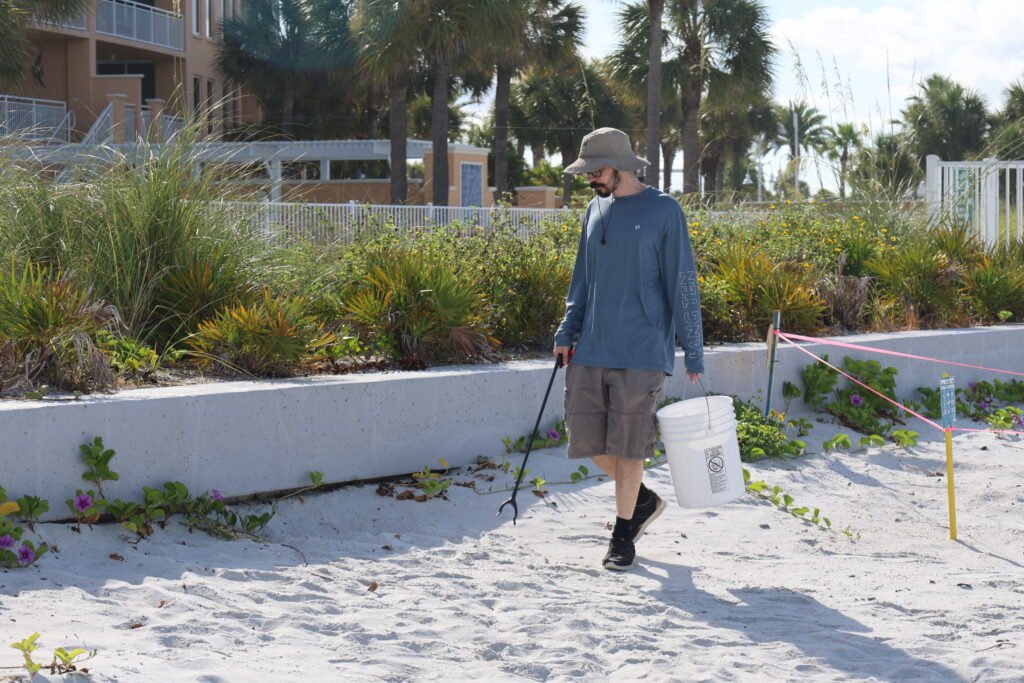 A volunteer walks along Madeira Beach carrying a bucket and litter picker during a beach cleanup, with coastal plants and palm trees in the background.