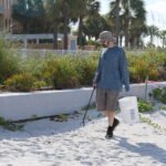 A volunteer walks along Madeira Beach carrying a bucket and litter picker during a beach cleanup, with coastal plants and palm trees in the background.